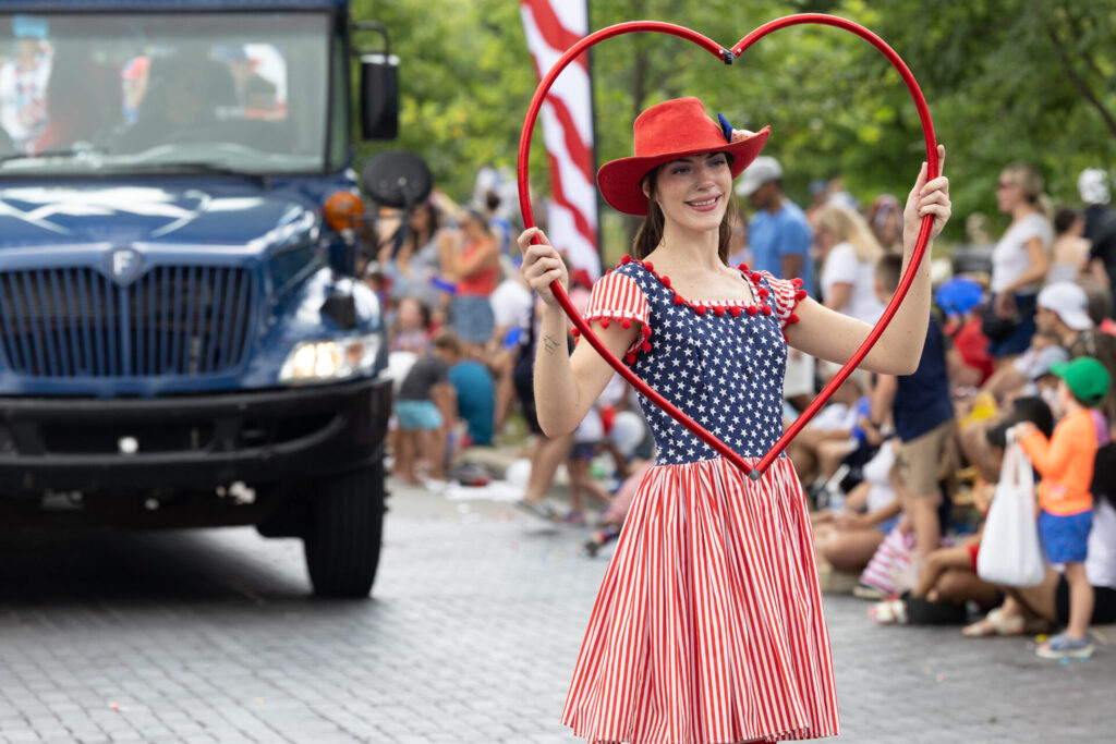 A smiling woman in a patriotic red, white, and blue dress and red hat holding a large red heart frame over her head during a parade with spectators and a blue truck in the background.