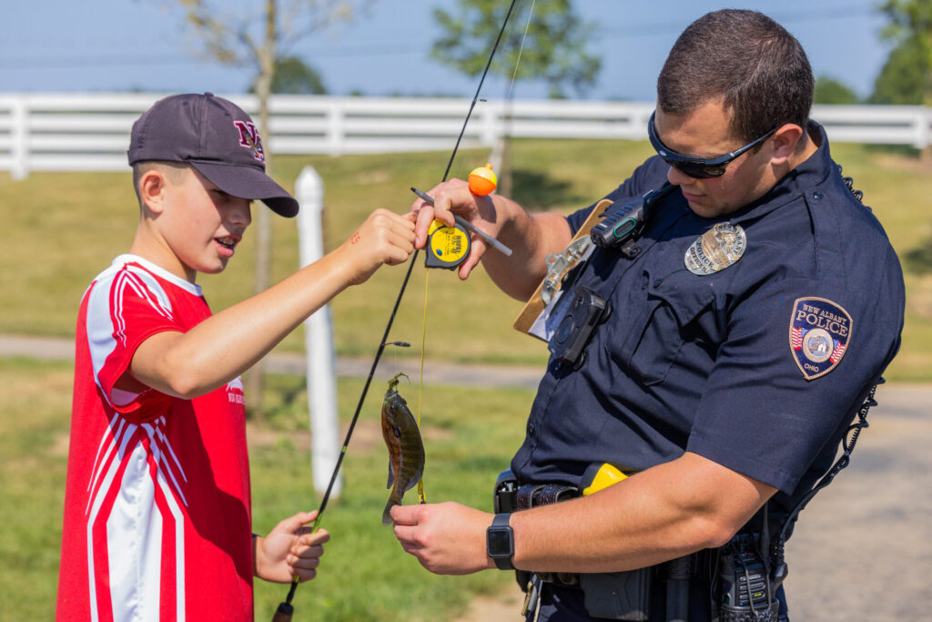Police officer helping a boy in a red shirt and cap hold up a fishing line with a small fish caught on the hook outdoors on a sunny day.