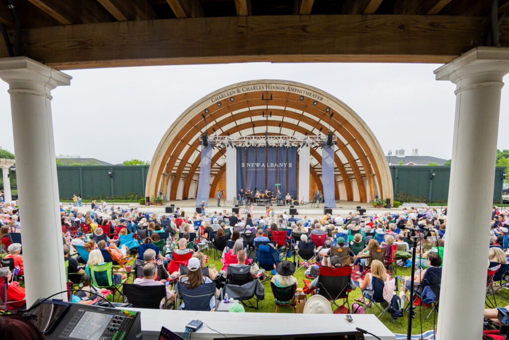 View from behind a sound mixing desk looking out over a large crowd seated on folding chairs facing the Charleen & Charles Hinson Amphitheater stage with performers and a New Albany banner in the background.