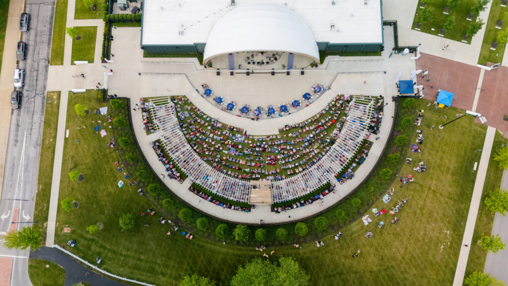 Aerial view of an outdoor amphitheater with a crowd seated on bleachers and lawn chairs facing a covered stage, surrounded by green grass and pathways.