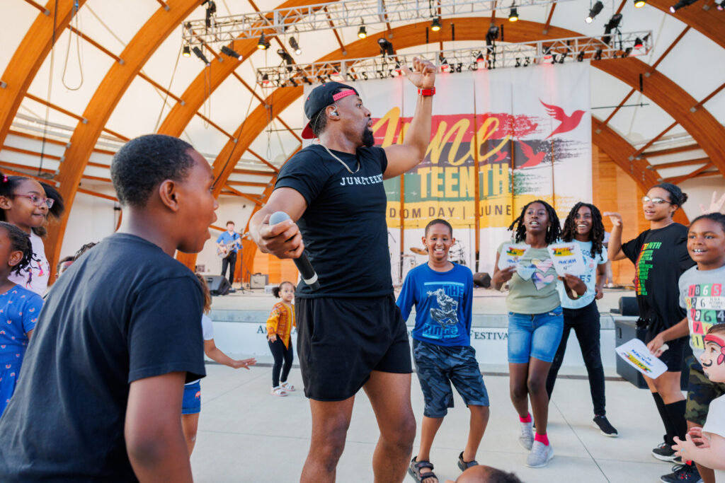 Man wearing a black Juneteenth t-shirt and cap holding a microphone, dancing with a group of excited children and teenagers at an outdoor event with a stage and a Juneteenth banner in the background.