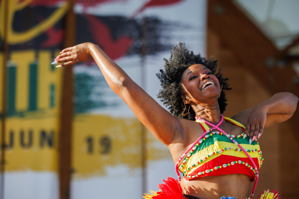 Joyful woman dancing outdoors wearing a colorful beaded top and hoop earrings, with her arms gracefully extended and an expressive smile on her face.