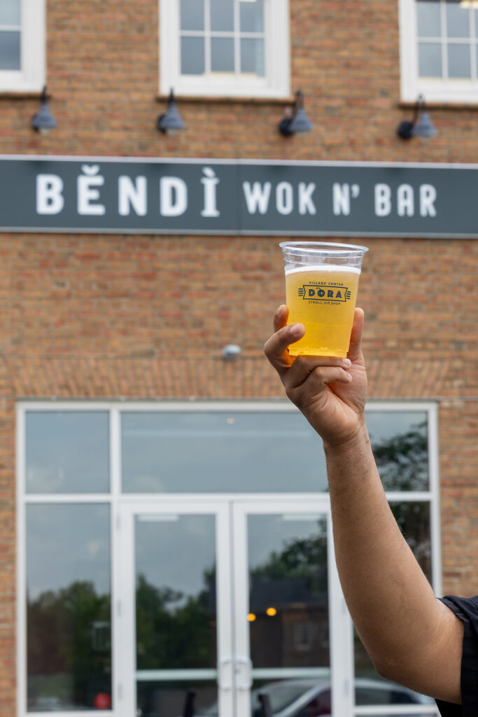 Hand holding up a plastic cup filled with beer in front of a brick building with a sign reading 'Bendi Wok N' Bar'