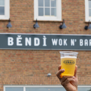 Hand holding up a plastic cup filled with beer in front of a brick building with a sign reading 'Bendi Wok N' Bar'