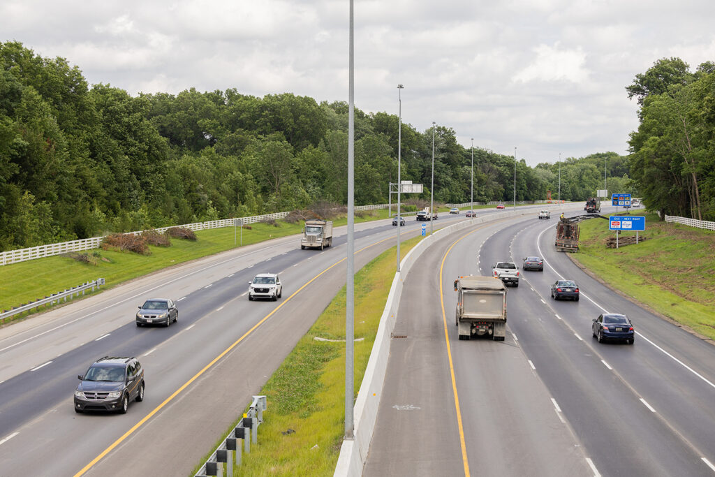 A divided highway with several cars and trucks driving in both directions, surrounded by green trees and grassy areas under a cloudy sky.