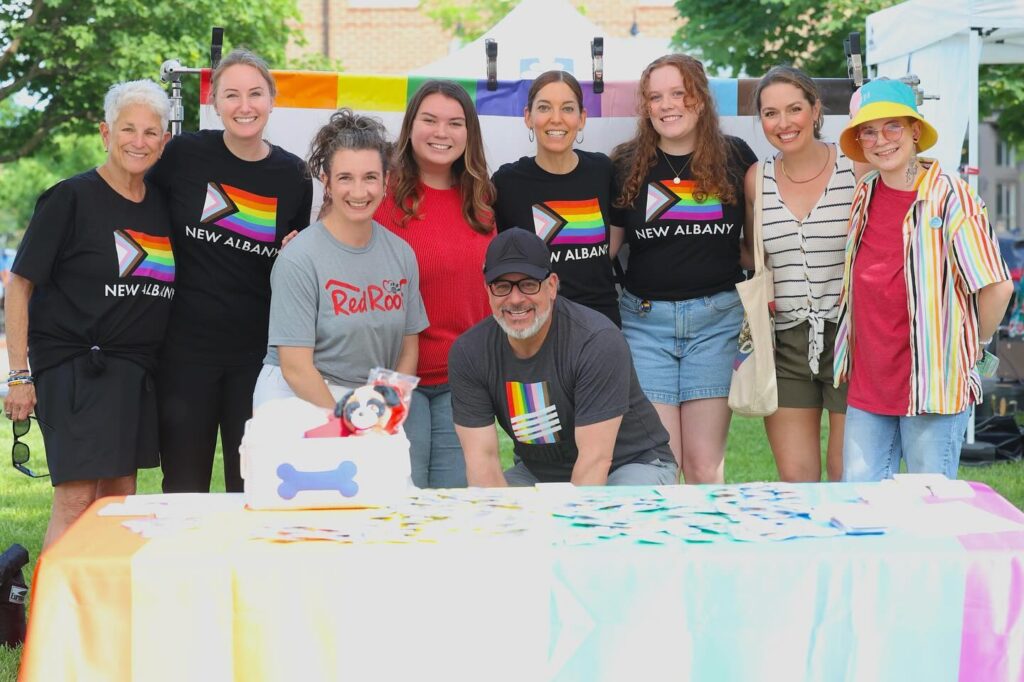 Group of nine people smiling and posing together outdoors behind a table covered with a rainbow pride flag, some wearing shirts with a progressive pride flag and New Albany text.