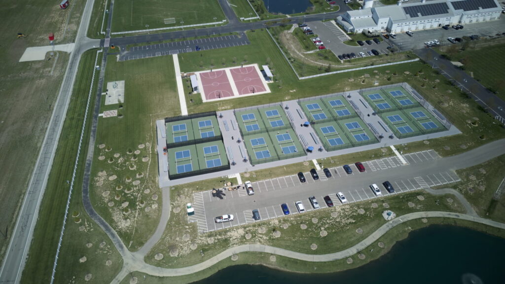Aerial view of a sports complex featuring multiple pickleball courts with blue and green surfaces, two red basketball courts, a parking lot with several cars, a road, grassy areas, and a nearby building with solar panels on the roof near a body of water.