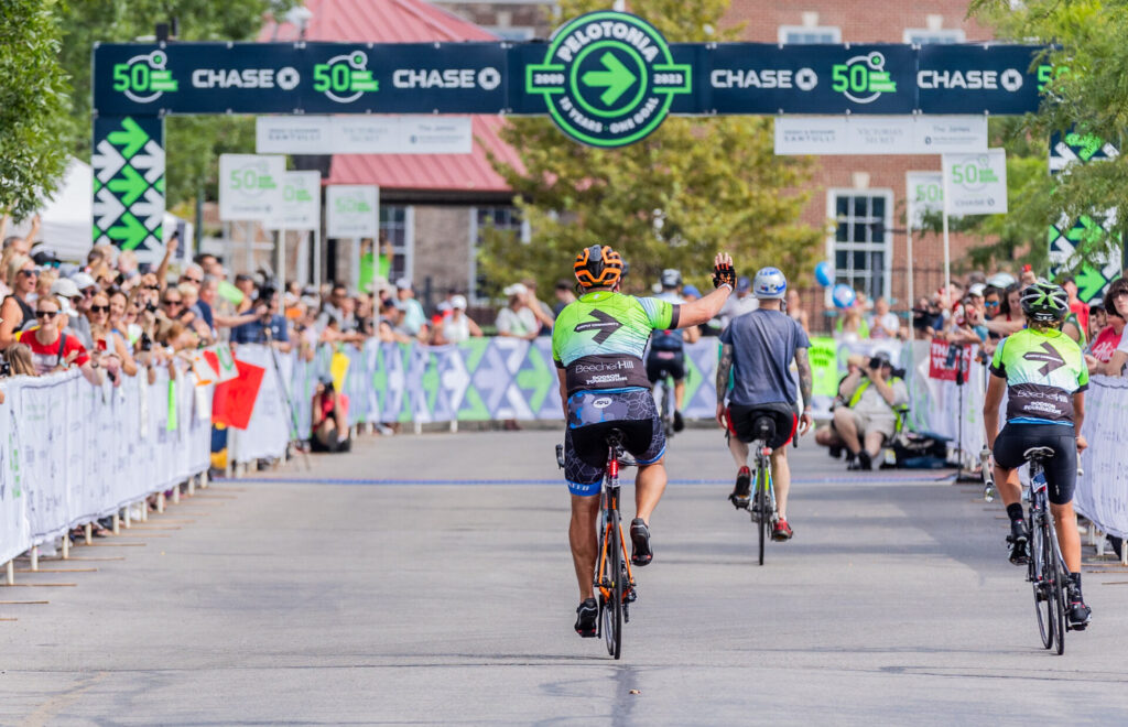 Cyclists riding towards a finish line banner that reads Pelotonia 2009-2023, with spectators cheering on both sides of the road during a cycling event.