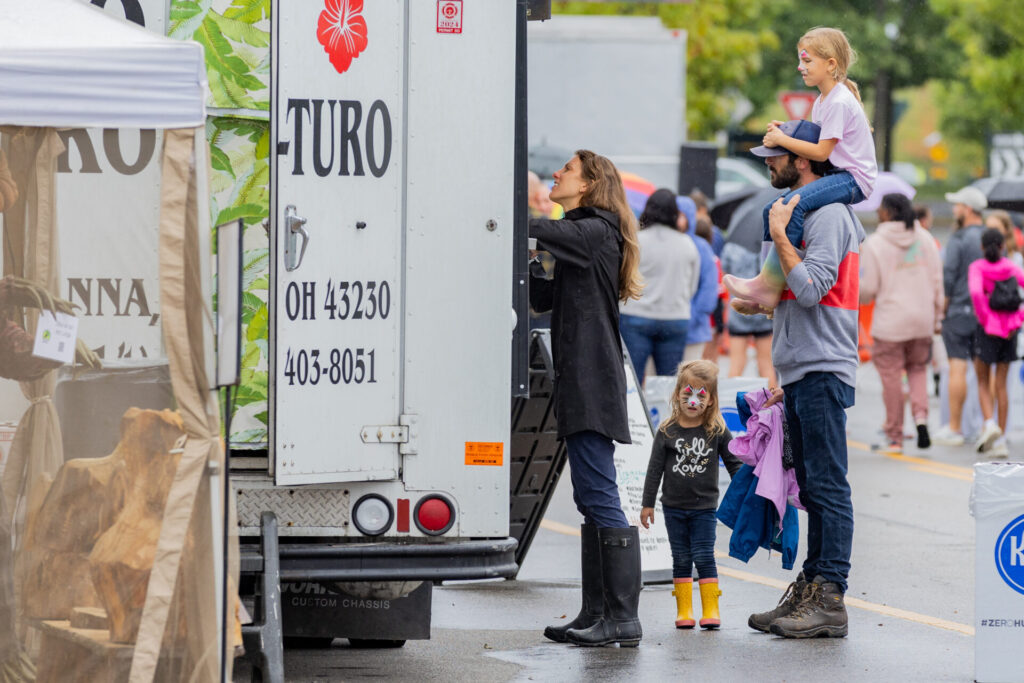 A woman in a black raincoat and boots orders from a food truck while a man carries a young girl on his shoulders and holds a jacket, with another young girl wearing face paint and yellow rain boots standing nearby, and a crowd in the background on a wet street.