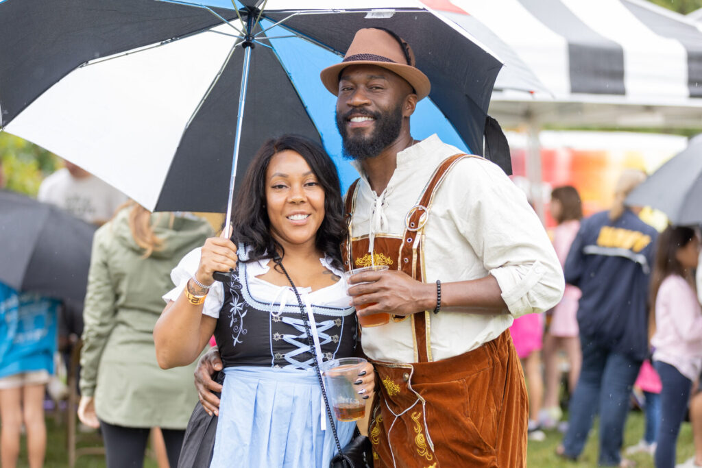 Smiling couple dressed in traditional German attire standing under a black and white umbrella at an outdoor event, each holding a plastic cup with a drink, with other people and tents visible in the background.