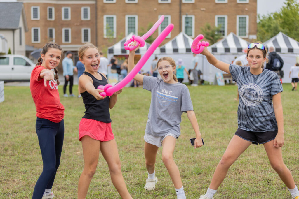 Four excited girls posing outdoors on grass holding pink balloon swords, with a background of striped tents and a brick building.