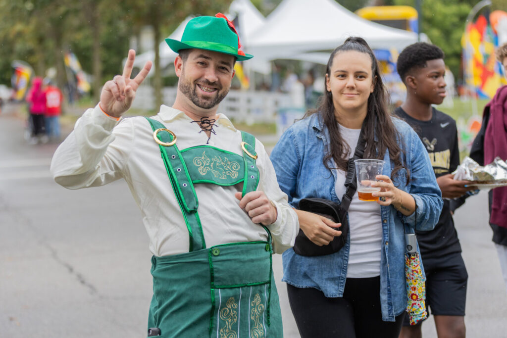 Man wearing traditional green Bavarian outfit with suspenders and hat making a peace sign, standing next to a woman in a denim jacket holding a plastic cup with a drink, with other people and event tents blurred in the background.