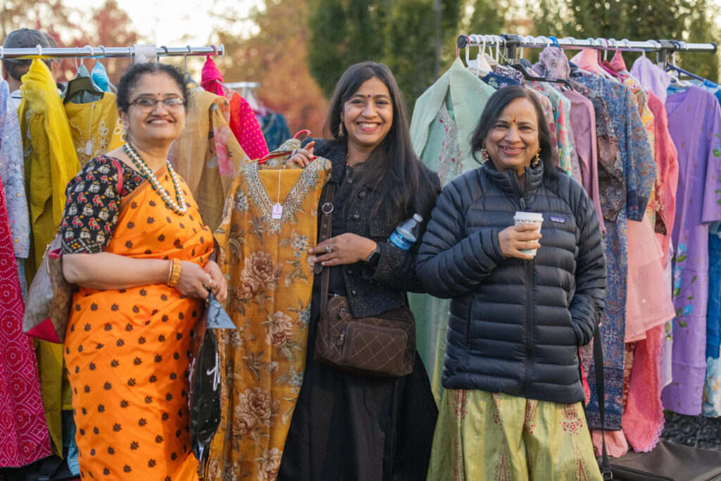 Three women smiling at a clothing stall outdoors with racks of colorful traditional garments behind them, one woman in an orange sari, one holding a floral-patterned mustard dress, and one in a black jacket holding a coffee cup.