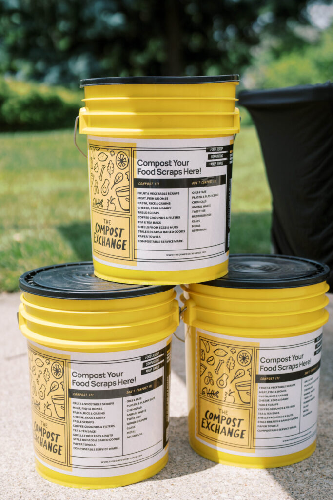 Three yellow compost buckets with black lids, labeled "The Compost Exchange" and instructions for composting food scraps, stacked outdoors on a concrete surface with grass and trees in the background.