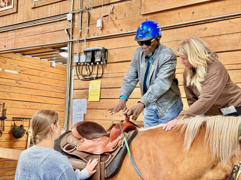 Two women assist a man wearing a blue helmet as he prepares to mount a brown horse with a leather saddle in a wooden indoor stable.