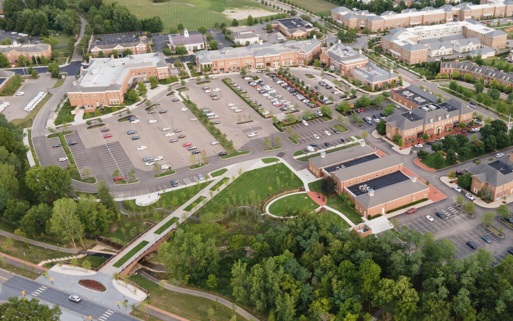 Aerial view of a suburban campus or office complex with multiple brick buildings, several parking lots with cars, green grassy areas, tree-lined pathways, and a small bridge over a wooded area near a street intersection.