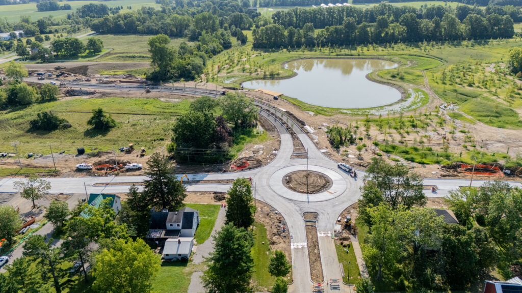 Aerial view of a rural area with a newly constructed roundabout surrounded by green fields, trees, and a small pond, with construction equipment and vehicles nearby.