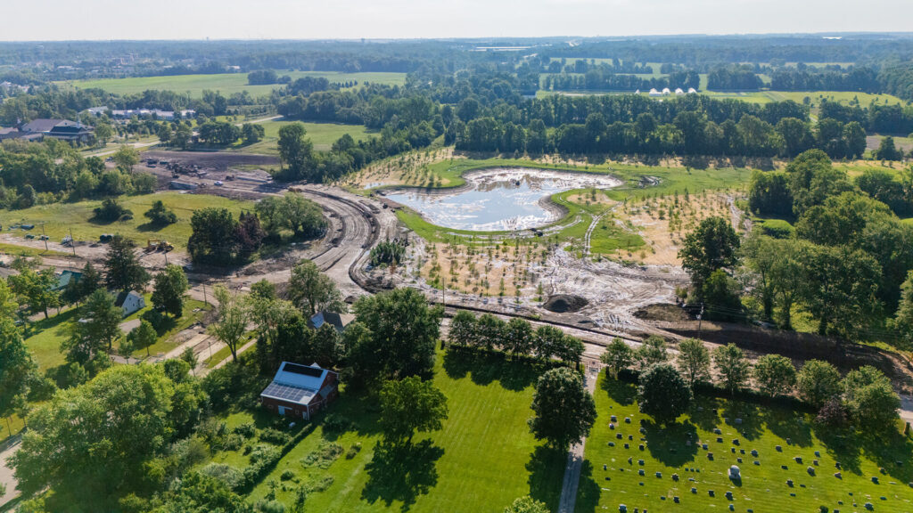Aerial view of a green rural landscape with a large muddy construction area surrounding a newly formed pond, scattered trees, houses, and open fields extending to the horizon under a clear sky.