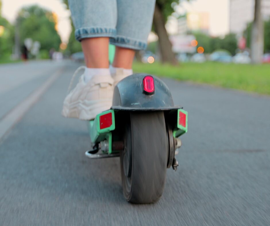 Close-up of a person wearing white sneakers and blue rolled-up jeans riding an electric scooter on a paved path with trees and buildings in the blurred background.