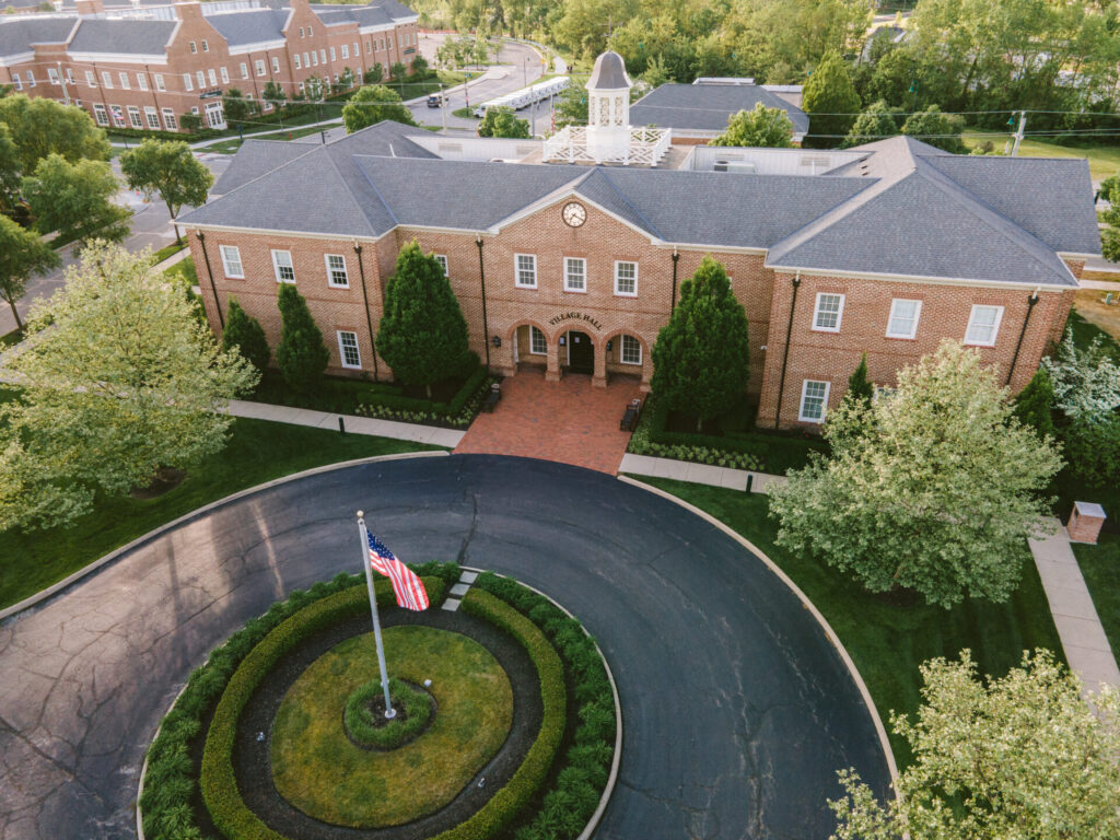 Aerial view of a brick village hall building with a clock and cupola on the roof, surrounded by green trees and a circular driveway featuring an American flag in the center.