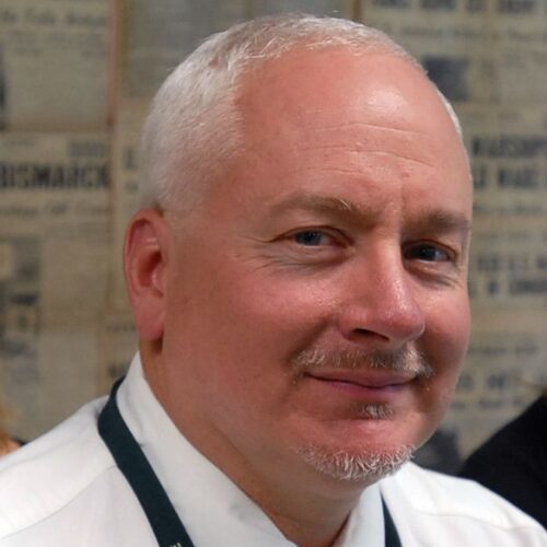 Middle-aged light-skinned man with short white hair and a trimmed goatee, giving a slight smile while wearing a white shirt and dark lanyard, with a blurred wall of printed text in the background.