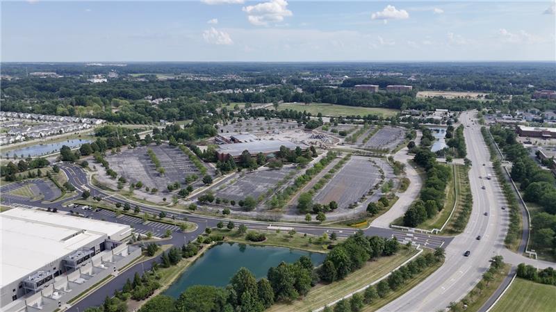 Aerial view of a suburban retail complex with large mostly empty parking lots, curving access roads and a small pond in the foreground, surrounded by trees, lawns and low-rise buildings under a partly cloudy sky.