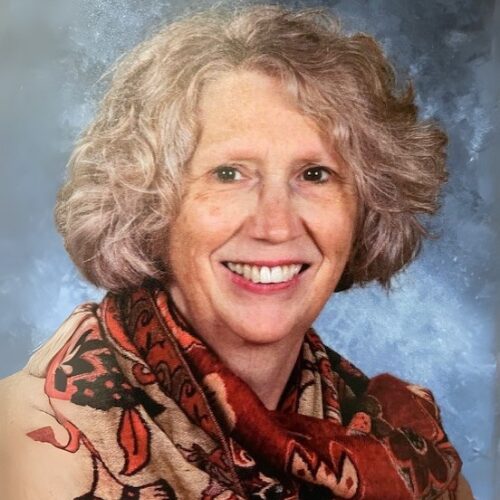 Smiling woman with short, curly gray hair wearing a red-and-beige patterned scarf, posed against a mottled blue-gray studio background.