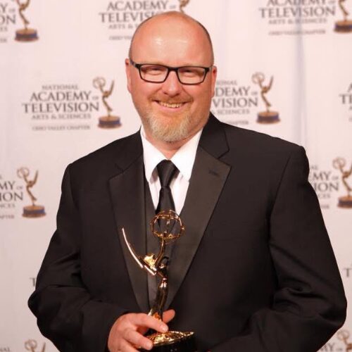 Smiling bald man with glasses and a trimmed beard wearing a black tuxedo and tie, holding an Emmy statuette in front of a National Academy of Television Arts & Sciences branded backdrop.