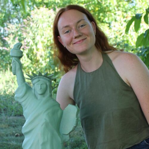 Red-haired woman in a sleeveless olive-green top smiling and leaning beside a small green Statue of Liberty replica outdoors with leafy trees in the background.