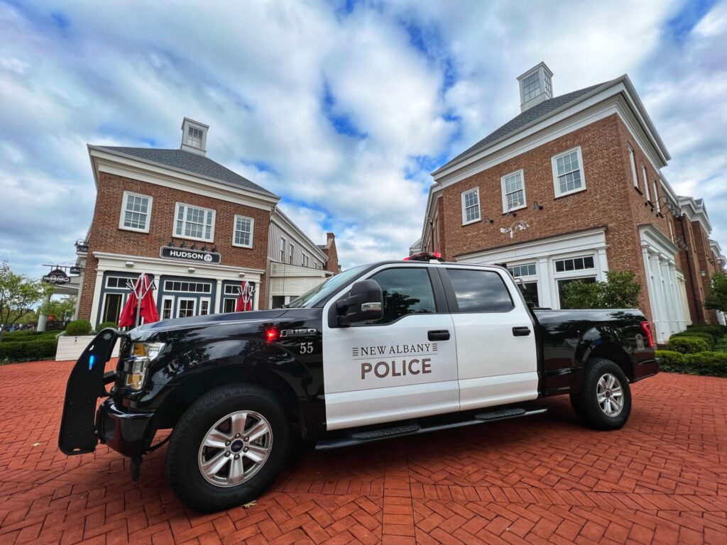 Black-and-white New Albany Police pickup truck with push bumper and lights parked on a red-brick plaza in front of two-story brick storefronts, one bearing a 'HUDSON' sign, under a cloudy blue-and-white sky.