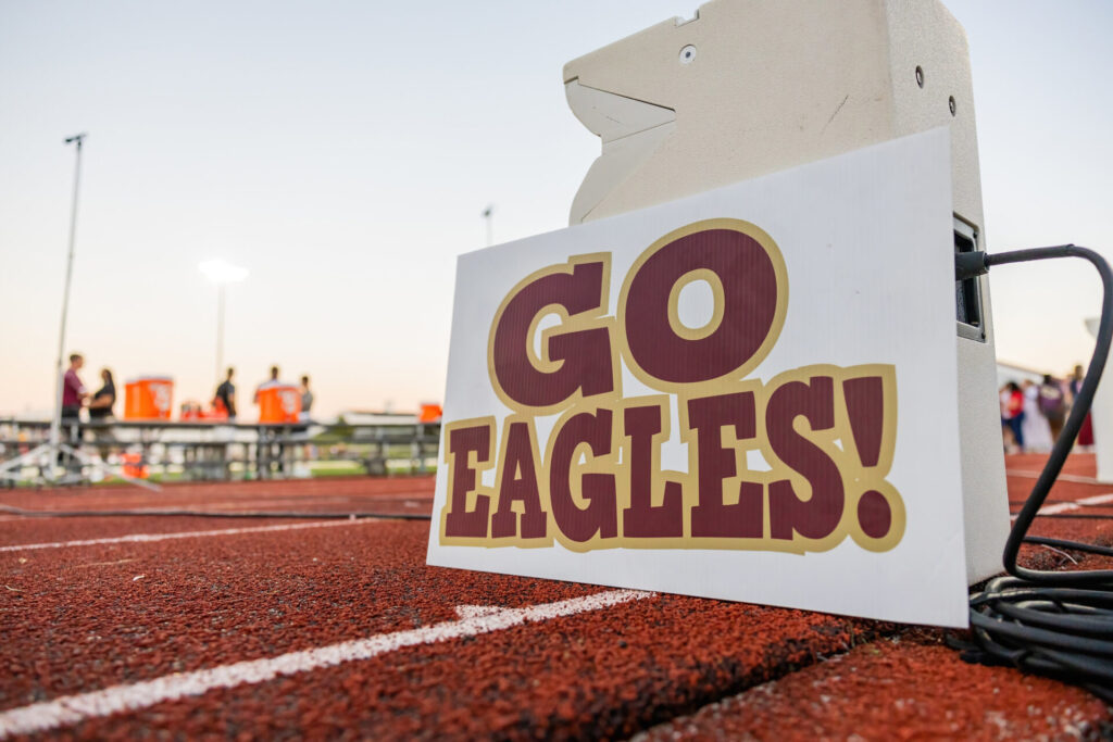 Sign reading GO EAGLES! in maroon and gold leaning against audio equipment on the edge of a red running track with blurred players, orange coolers and stadium lights in the background.