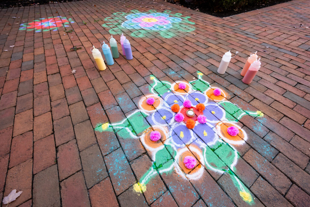 Colorful rangoli pattern of purple, orange, green and white powdered chalk with pink pom-pom accents and a central tealight on a red brick walkway, with squeeze bottles of paint nearby and additional chalk designs visible in the background.