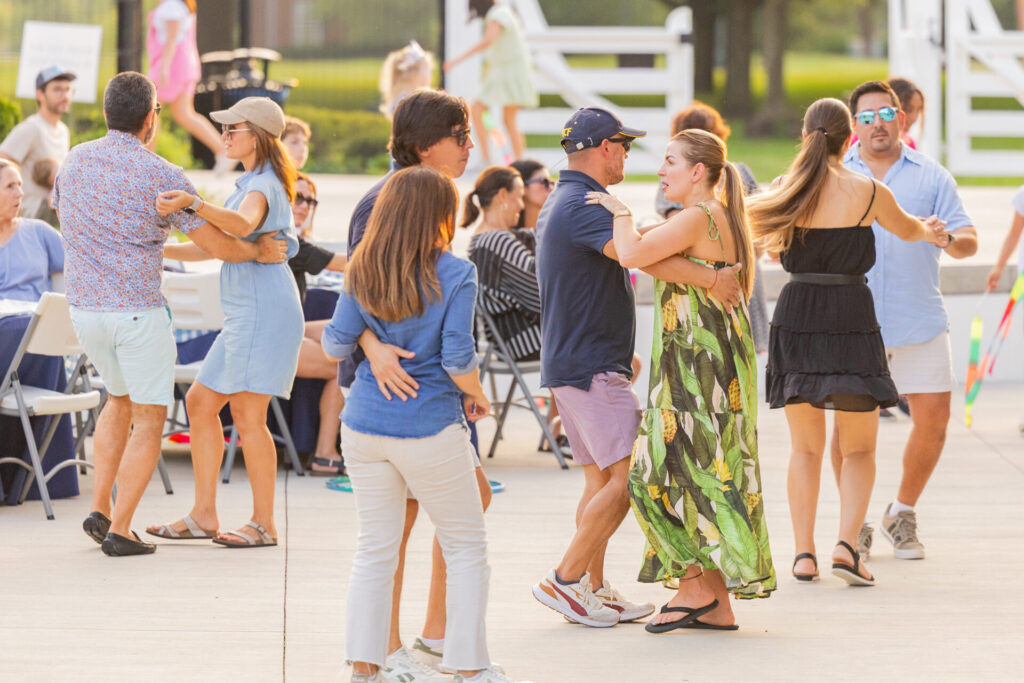 Several couples dance on a sunny outdoor patio, centered on a woman in a green pineapple-print maxi dress dancing with a man in a navy cap and shorts, with other casually dressed partners and folding chairs and tables in the background.