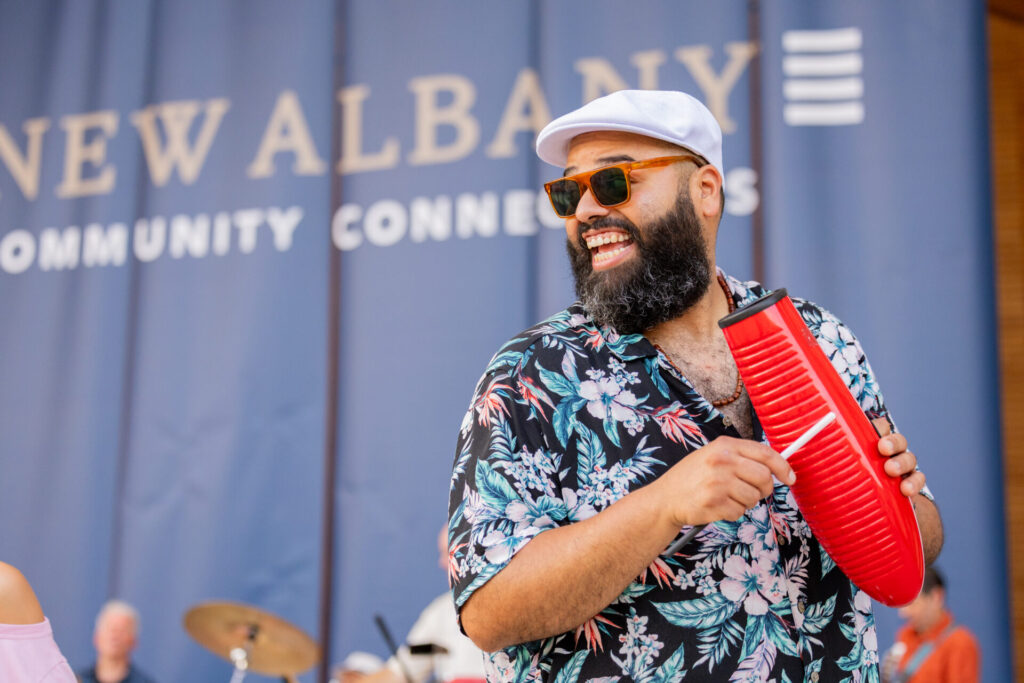 Bearded man in a white cap, sunglasses and a floral shirt smiles while scraping a red güiro with a stick onstage in front of a blue "New Albany Community" banner.