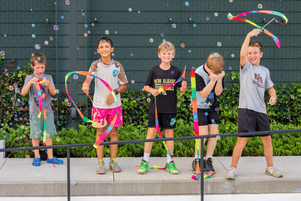 Five boys stand on a low concrete ledge in front of a green fence and shrubs, smiling and playing with long rainbow ribbons and soap bubbles — one waves a ribbon high in the air, another examines a ribbon, and the others laugh as bubbles float around them.