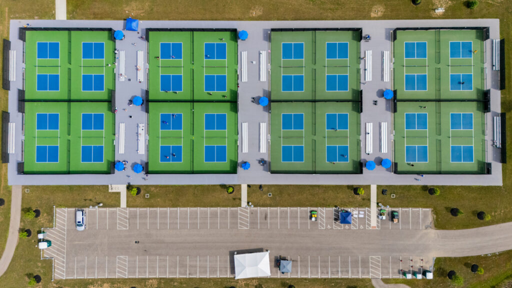 Aerial view of eight tennis courts arranged in two rows with blue and green surfaces, surrounded by green grass, white benches, blue umbrellas, and a mostly empty parking lot at the bottom of the image.