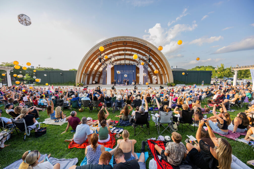 Large crowd on blankets and folding chairs fills a grassy lawn watching performers on a wooden, semi‑circular outdoor amphitheater as yellow and clear balloons float overhead against a blue sky.