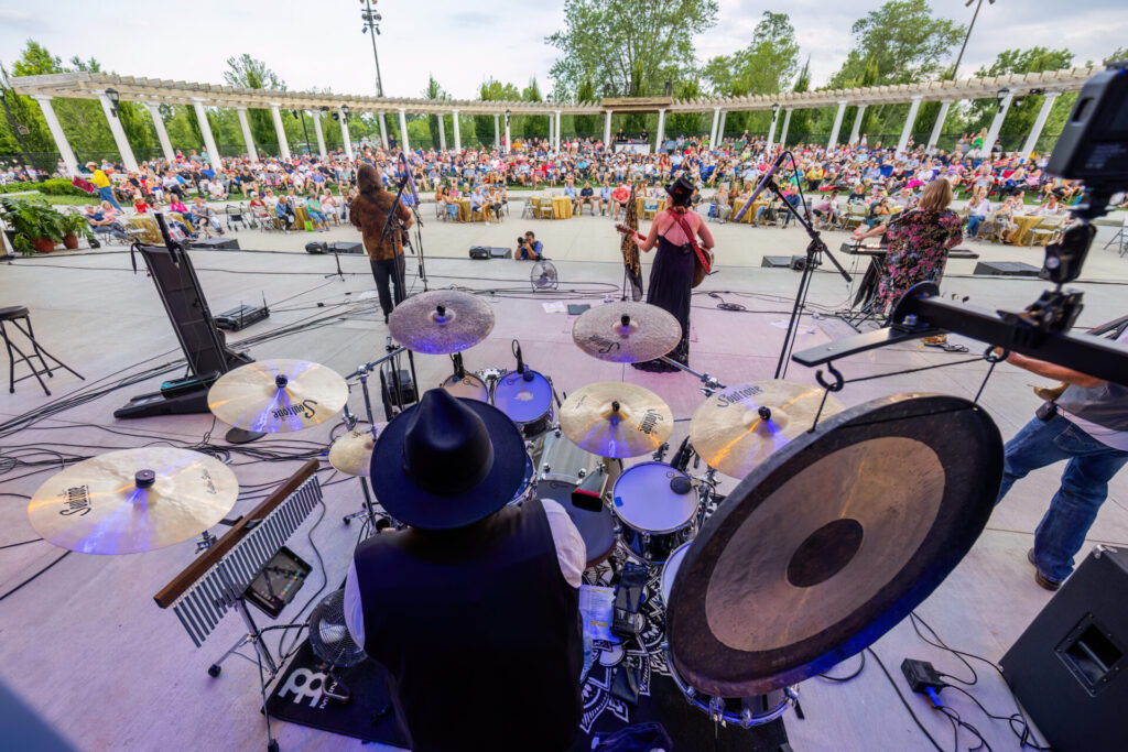 View from behind a drummer in a black hat showing drum kit and cymbals in the foreground as a band performs on an outdoor amphitheater stage before a large seated audience under a white pergola, with trees in the background.