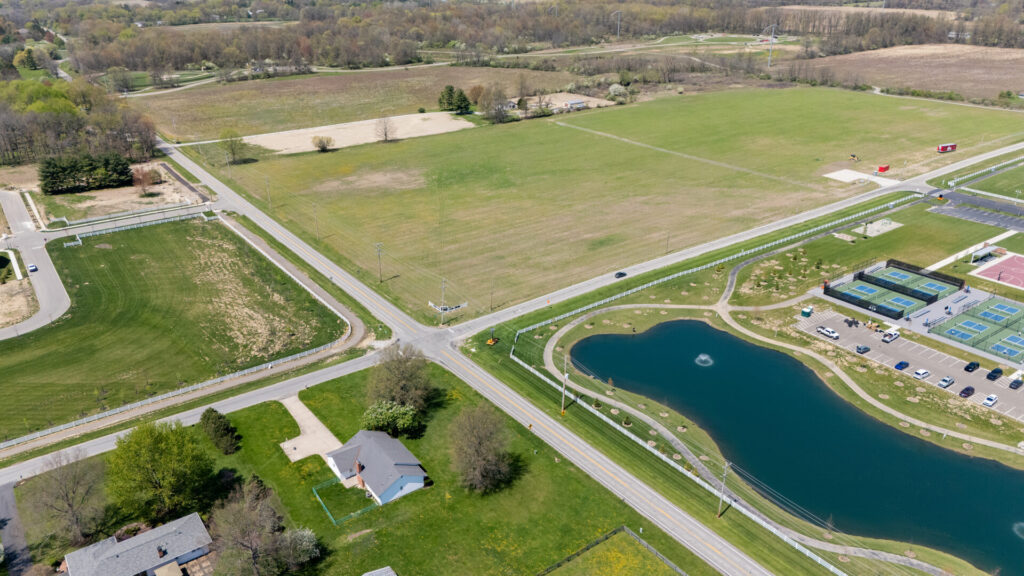 Aerial view of a rural intersection with open fields, a pond with a fountain, tennis courts, and scattered houses surrounded by greenery and trees.