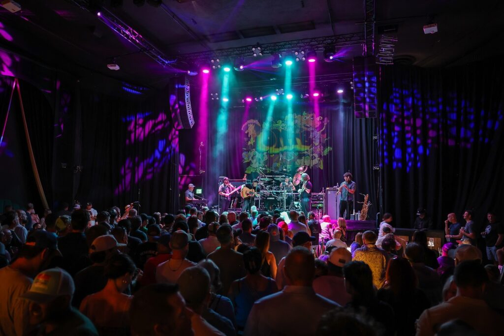 Packed indoor concert venue with a live band—drummer, guitarist, keyboardist and saxophonist—performing on a stage under teal, purple and magenta spotlights and fog beams while a dense crowd watches and a colorful decorative backdrop hangs behind the band.