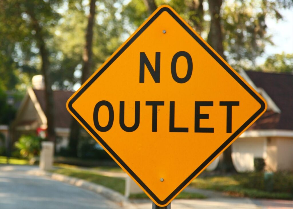 Yellow diamond-shaped traffic sign with black text reading No Outlet, set in a suburban neighborhood with trees and houses in the background.