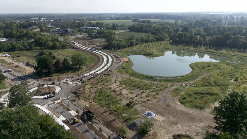 Aerial view of a construction site showing a newly paved curved road and roundabout beside an irregularly shaped pond, surrounded by planted trees, grassy fields, and construction equipment.