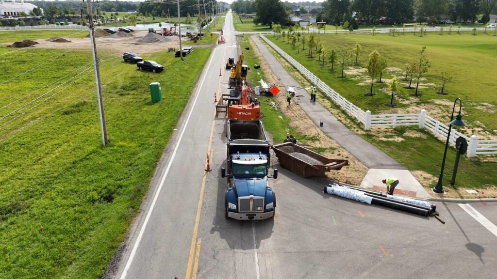 Aerial view of a roadside construction site with a blue dump truck, an orange Hitachi excavator, construction materials, and workers in safety vests near a paved path and grassy fields with white fences.
