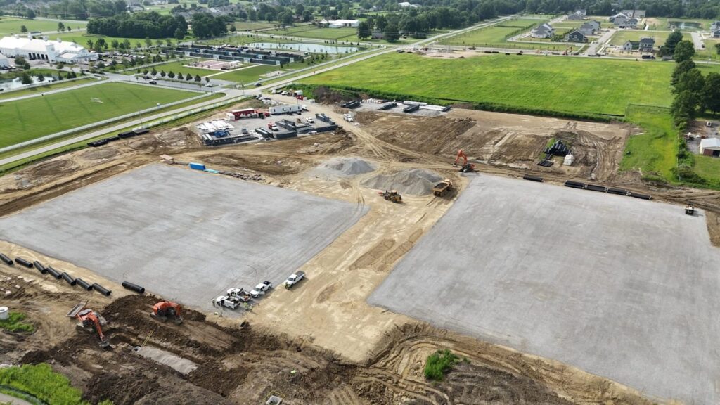 Aerial view of a large construction site with cleared and leveled ground, piles of gravel, construction vehicles including excavators and trucks, and stacks of black pipes, bordered by green fields and residential houses in the background.