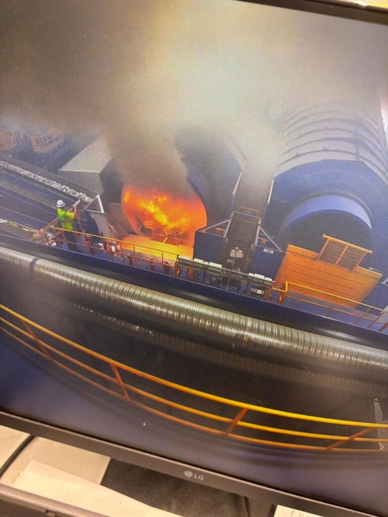 A worker in a high-visibility jacket and hard hat stands on a walkway as bright orange flames and thick smoke pour from a large industrial rotary drum, with yellow safety railings and large cylindrical rollers in the foreground.
