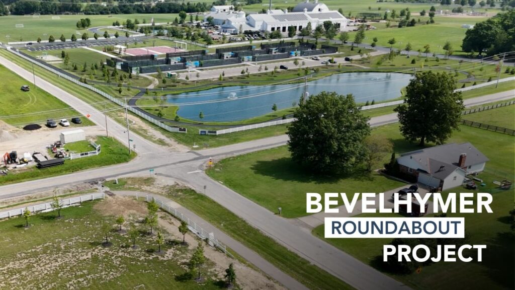 Aerial view of a roundabout project near a pond with fountains, adjacent to a large white building, sports courts, parking lots, roads, green fields, and trees, with the text 'BEVELHYMER ROUNDABOUT PROJECT' overlayed on the right side.