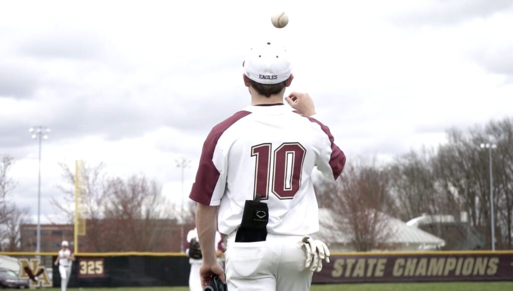 Baseball player in a maroon-and-white uniform with number 10 and "EAGLES" on his cap, seen from behind as he tosses a ball overhead on a field with an outfield fence banner reading STATE CHAMPIONS.