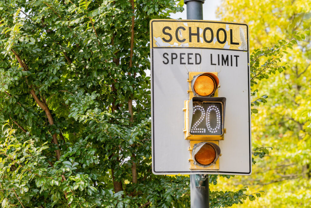 School zone speed limit sign on a pole with two amber lights and an illuminated '20' display, set against leafy green trees.
