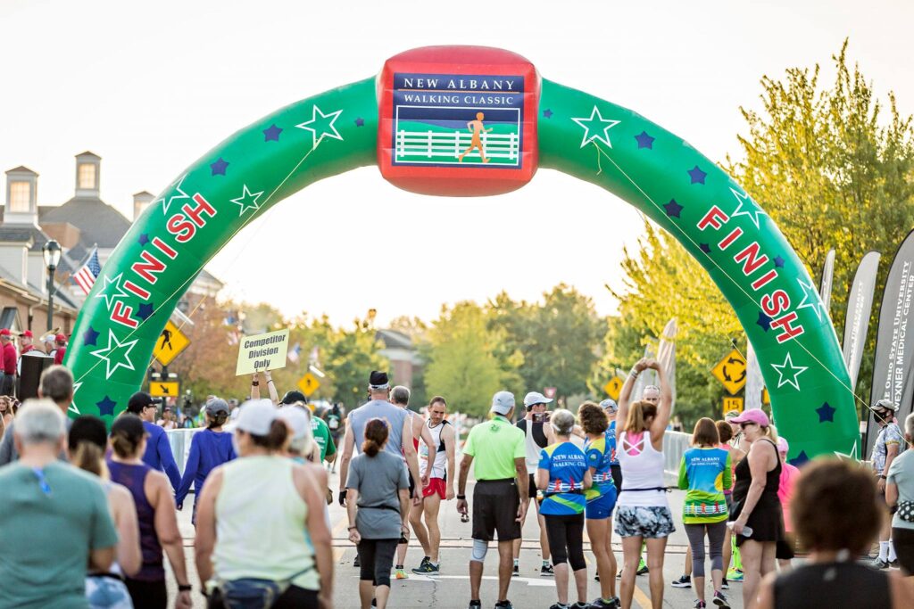 Crowd of walkers and runners pass under a large green inflatable arch reading FINISH and New Albany Walking Classic on a tree-lined street, while a volunteer holds a Competitor Division Only sign.