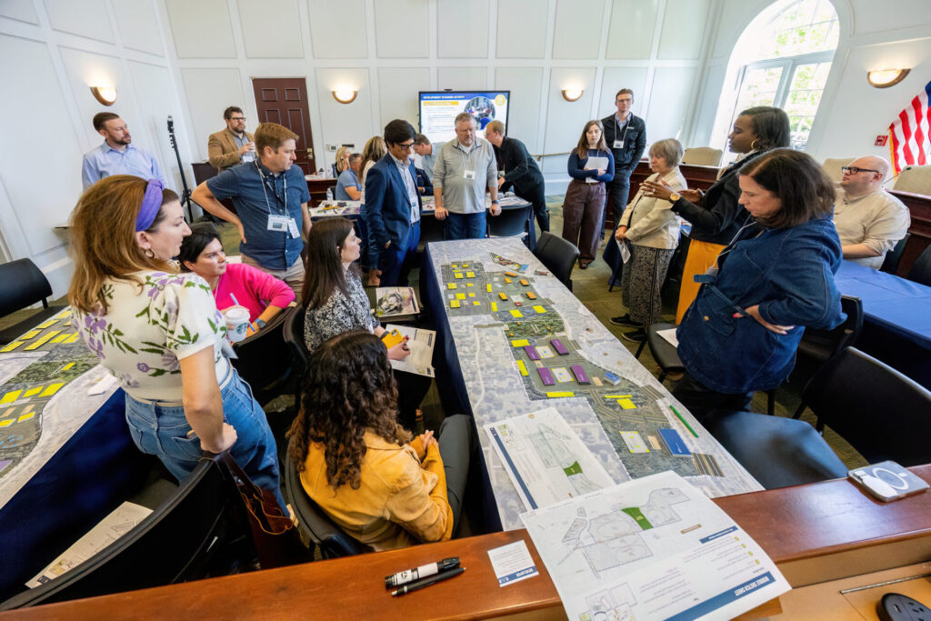A diverse group of people stands and sits around a long table covered with large printed maps, color-coded sticky notes and planning documents in a bright meeting room while one person gestures and others listen and examine the layouts.